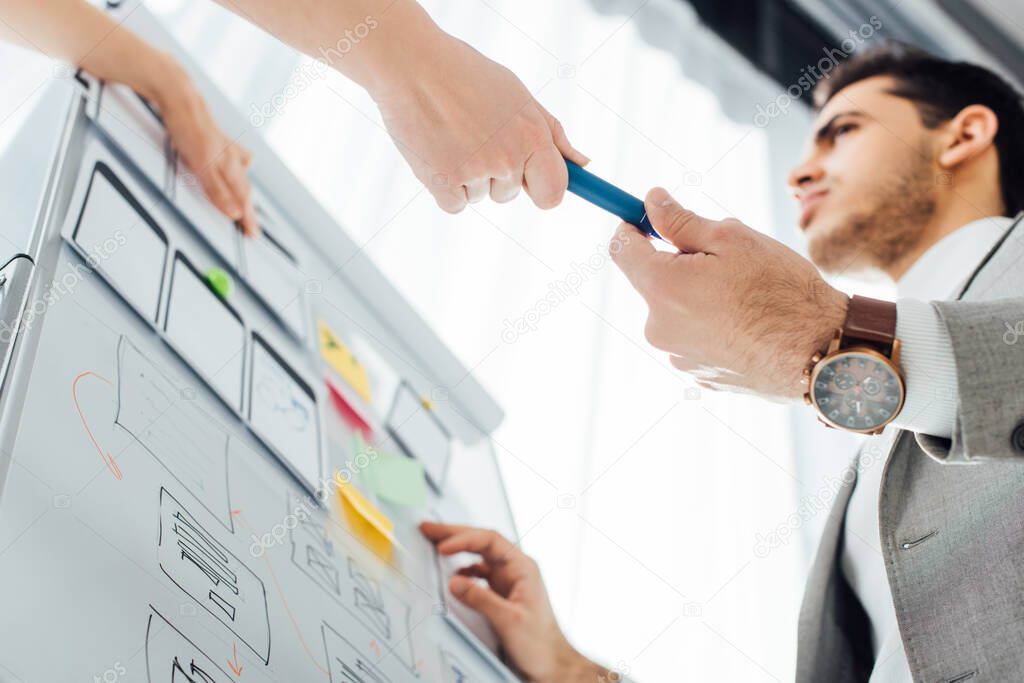 Low angle view of ux designer giving marker to colleague near layouts on whiteboard in office 