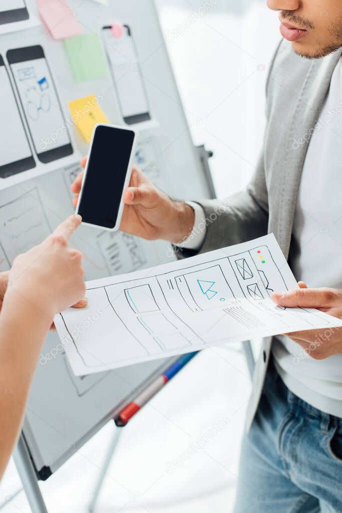 Cropped view of designer pointing on smartphone near colleague with templates of ux design near whiteboard in office 