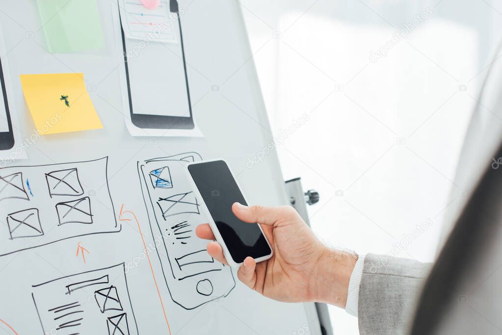 Selective focus of ux designer using smartphone near sketches and layouts on whiteboard in office 