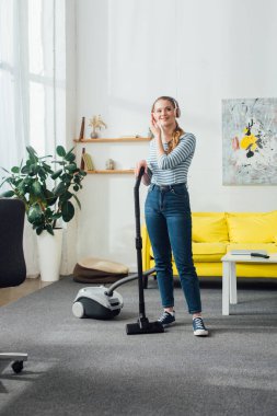 Oturma odasında elektrikli süpürgenin yanında müzik dinlerken gülümseyen kulaklı güzel bir kız.