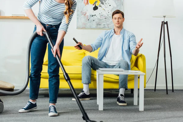 Frau Putzt Teppich Neben Überraschtem Freund Mit Fernbedienung Auf Couch — Stockfoto