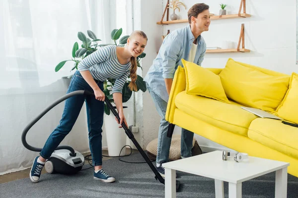 Glimlachende Man Tillen Bank Buurt Vriendin Met Stofzuiger Woonkamer — Stockfoto