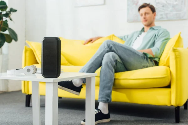 Selective focus of wireless speaker and headphones on coffee table and man sitting on couch in living room