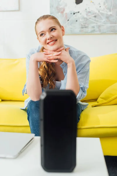 紧握着双手坐在靠近无线扬声器的沙发上和咖啡桌上笔记本电脑的微笑女性的选择性聚焦 — 图库照片