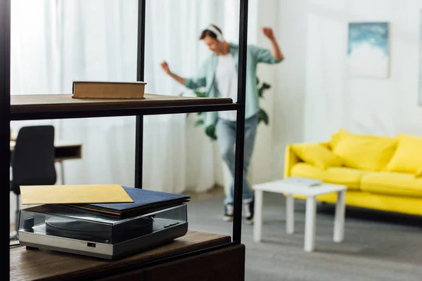 Selective focus of book and record player on shelves of cabinet and man in headphones dancing at home