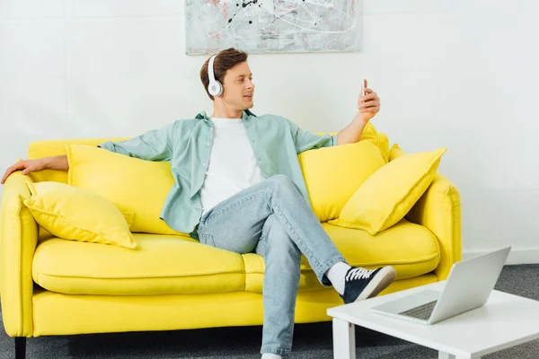 Schöner Mann Mit Kopfhörer Und Smartphone Auf Couch Neben Laptop — Stockfoto