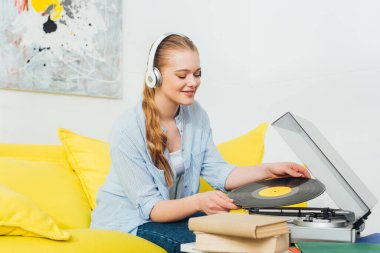 Kulaklıklı gülümseyen kadın pikabın yanında vinil plak ve sehpanın üzerinde kitaplar tutuyor. 