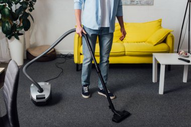 Oturma odasında halı temizlerken elektrikli süpürge kullanan bir adamın görüntüsü.