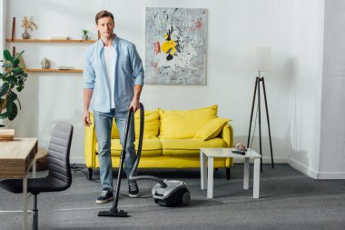 Oturma odasında elektrikli süpürge kullanırken kameraya bakan yakışıklı adam.