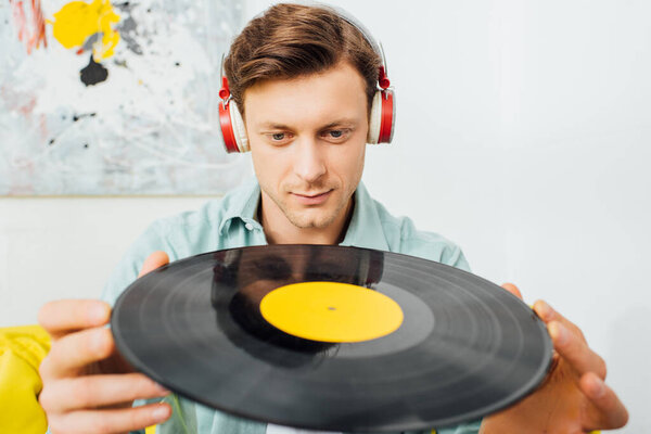 Selective focus of handsome man in headphones holding vinyl record in living room