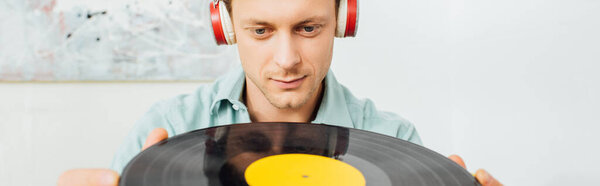 Panoramic shot of handsome man in headphones holding vinyl record in living room