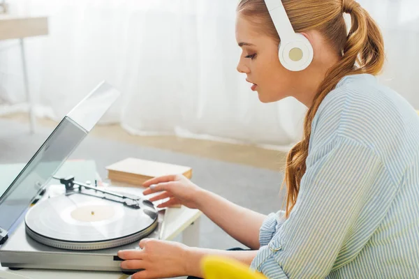 Side view of beautiful woman in headphones using record player near books on coffee table at home