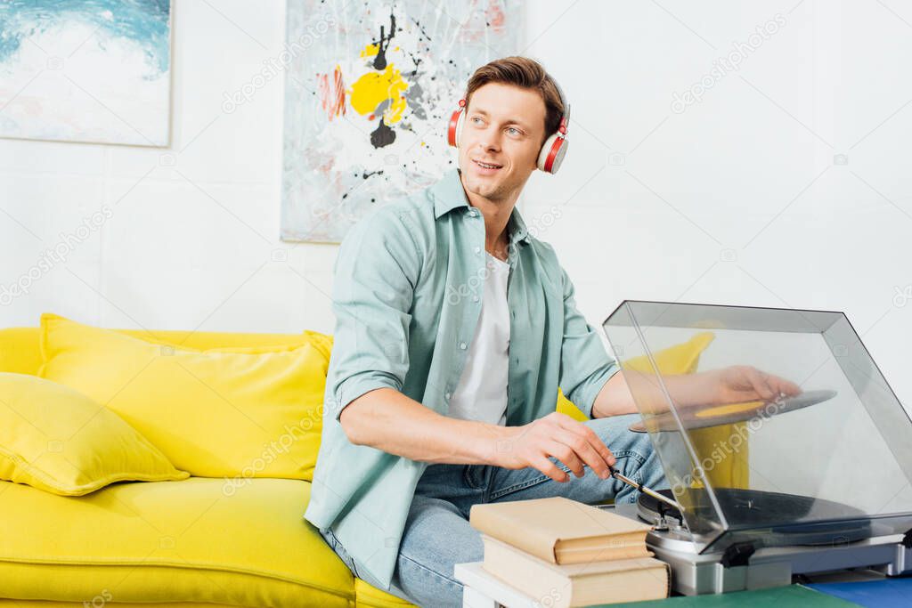 Smiling man in headphones looking away while using record player near books on coffee table