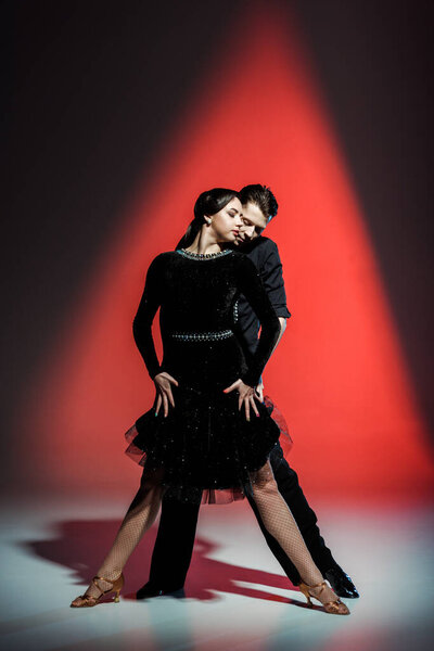 elegant young couple of ballroom dancers in black outfits dancing in red light