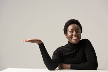 Siyah balıkçı yaka kazaklı neşeli Afro-Amerikan kadın masada oturuyor ve gri üzerinde izole edilmiş bir şey gösteriyor.