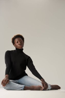Siyah balıkçı yaka yakalı güzel Afro-Amerikalı kız gri kübün üzerinde yalnız başına oturuyor.