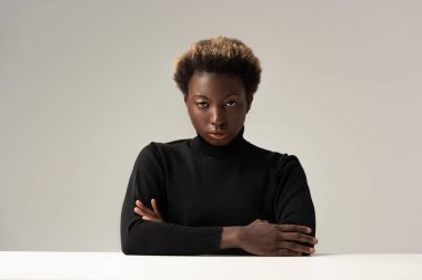 Siyah balıkçı yakalı Afro kadın masada oturuyor kolları çapraz ve gri renkte.