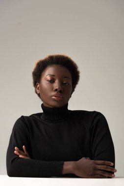 Siyahi balıkçı yakalı Afro-Amerikan kadın masada oturuyor kolları çapraz ve gri renkte.