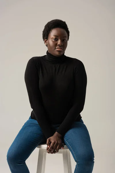 african american girl in jeans and black turtleneck sitting on stool isolated on grey