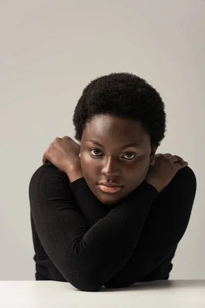 african american woman in bad mood sitting at table isolated on grey