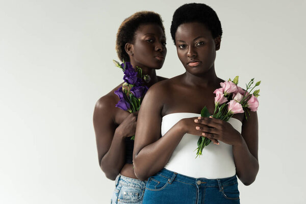 african american women in jeans and tops holding flowers isolated on grey
