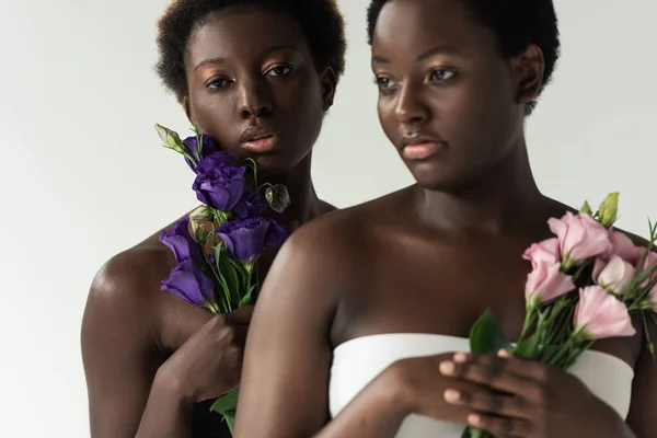 attractive african american women in tops holding pink and purple flowers isolated on grey