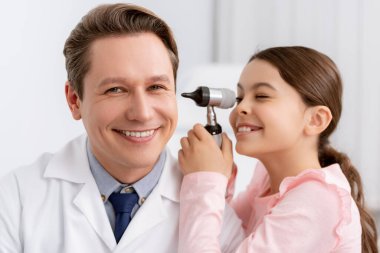 Mutlu çocuk otoskoplu gülümseyen doktorun kulağını inceliyor.