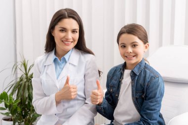 Mutlu doktor ve çocuk kameraya gülümserken başparmaklarını gösteriyorlar.