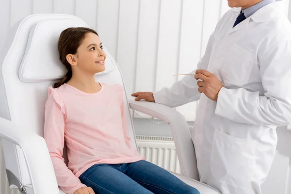 站在坐在医疗椅上笑着的小孩旁边时 耳鼻咽学家拿着喉头吐痰的剪影 — 图库照片