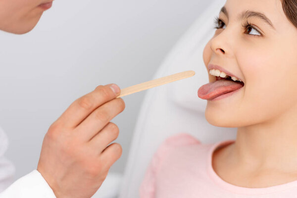 cropped view of otolaryngologist holding tongue depressor near kid sticking out tongue 