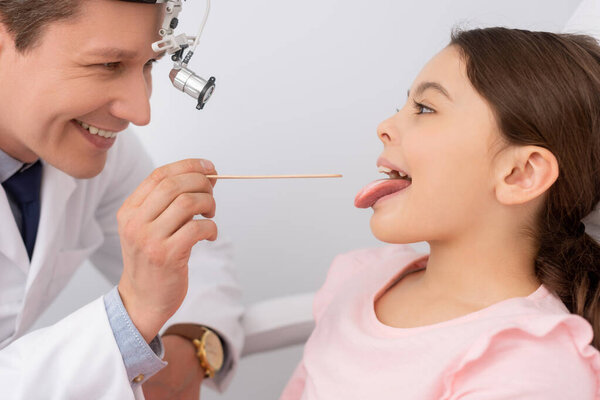 smiling ent physician examining throat of cute child with tongue depressor