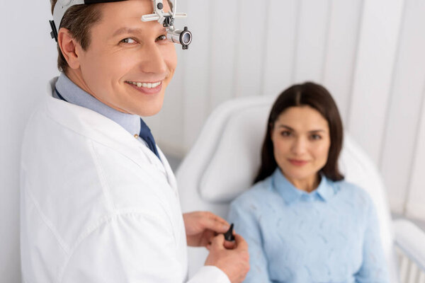 selective focus of smiling otolaryngologist in ent headlight holding ear speculum near smiling woman sitting in medical chair