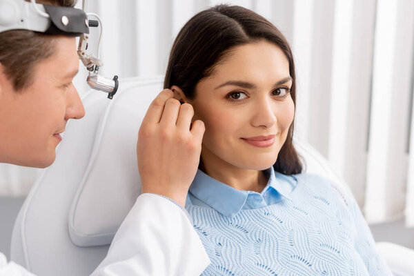 smiling woman looking at camera while otolaryngologist examining her ear 