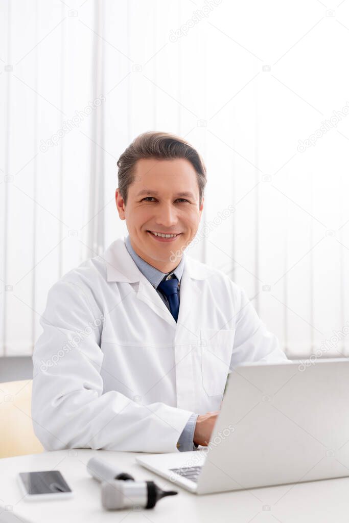 smiling otolaryngologist looking at camera while using laptop at workplace