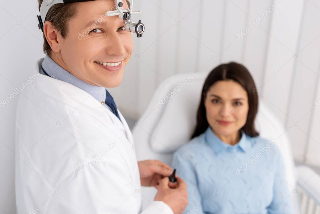 selective focus of smiling otolaryngologist in ent headlight holding ear speculum near smiling woman sitting in medical chair