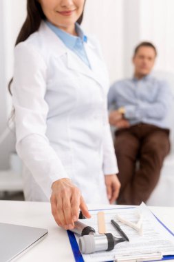Doktorun otoskop alıp gülümsemesi ve hastanın arka planda tıbbi sandalyede oturması.