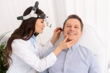 attractive smiling otolaryngologist in ent headlight examining ear of cheerful man  clipart