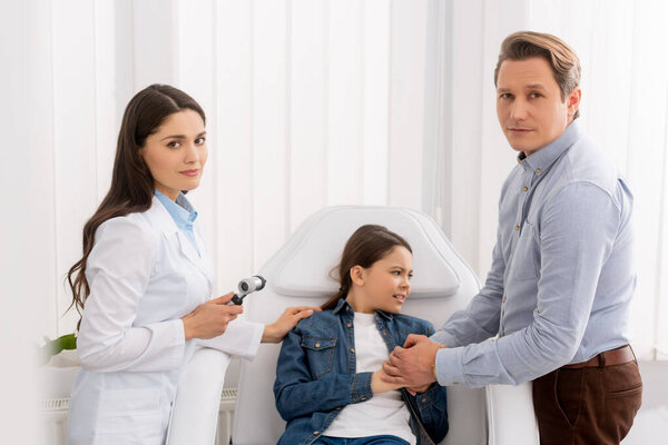 father holding hands of worried daughter sitting in medical chair near ent physician with otoscope