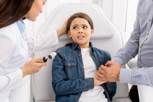 cropped view of father touching hands of daughter near otolaryngologist with otoscope