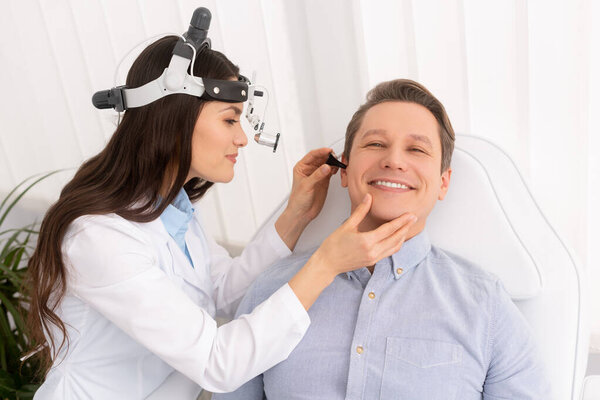 attractive smiling otolaryngologist in ent headlight examining ear of cheerful man 