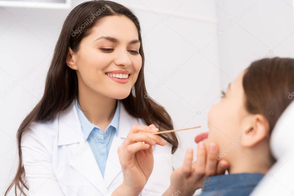 selective focus of smiling otolaryngologist examining throat of kid with tongue depressor