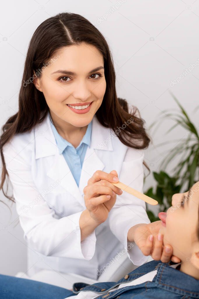 smiling otolaryngologist looking at camera while examining throat of child with tongue depressor