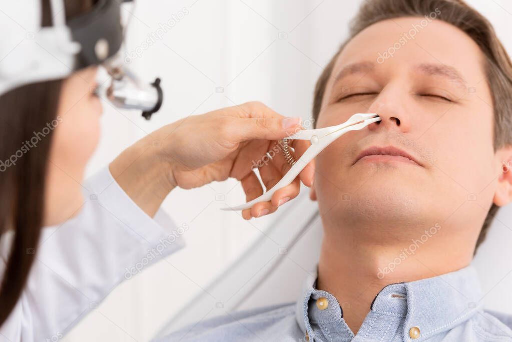 cropped view of otolaryngologist examining nose of handsome man with nasal speculum