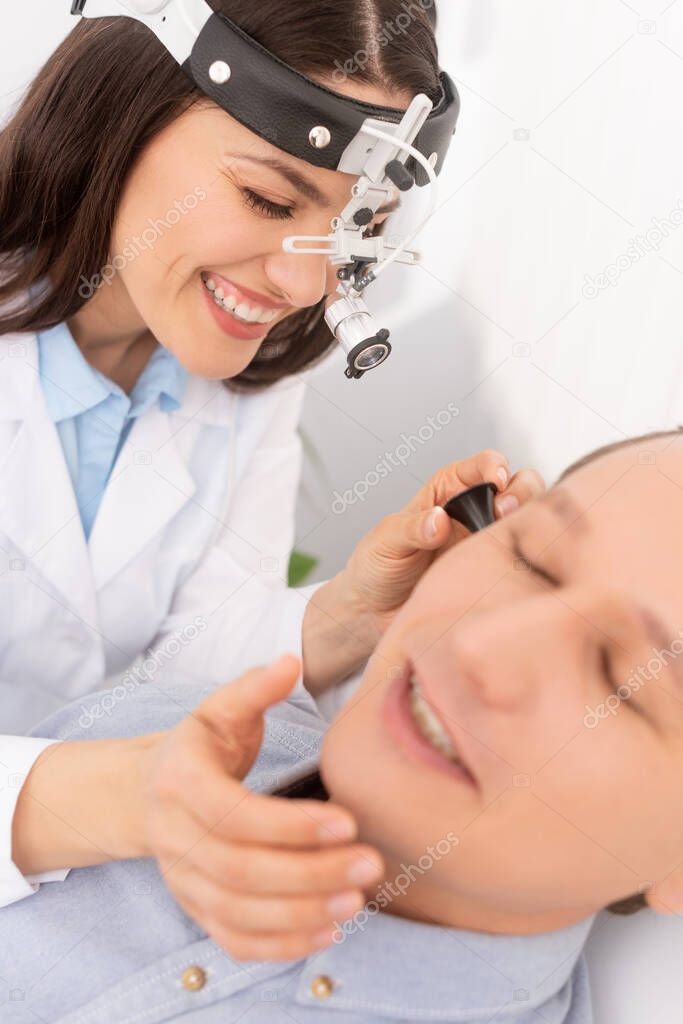 selective focus of attractive otolaryngologist in ent headlight examining ear of smiling man 