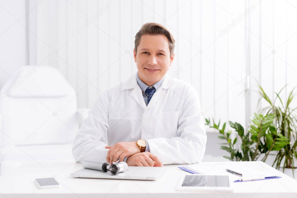 smiling ent physician smiling while sitting at workplace near otoscope