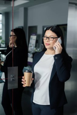 Çekici Asyalı iş kadını elinde kağıt bardakla akıllı telefondan konuşuyor. 
