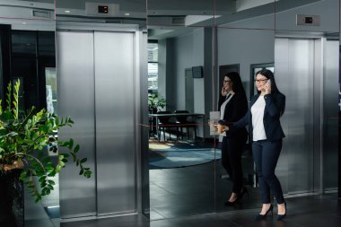 gülümseyen Asyalı iş kadını asansörün düğmesine basıyor ve akıllı telefondan konuşuyor. 
