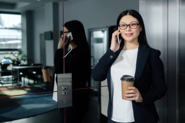 gülümseyen Asyalı iş kadını elinde kağıt bardakla akıllı telefondan konuşuyor. 