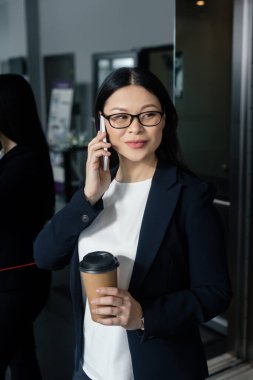gülümseyen Asyalı iş kadını elinde kağıt bardakla akıllı telefondan konuşuyor. 