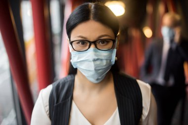 Resmi giyinmiş, tıbbi maskeli Asyalı bir iş kadını kameraya bakıyor. 